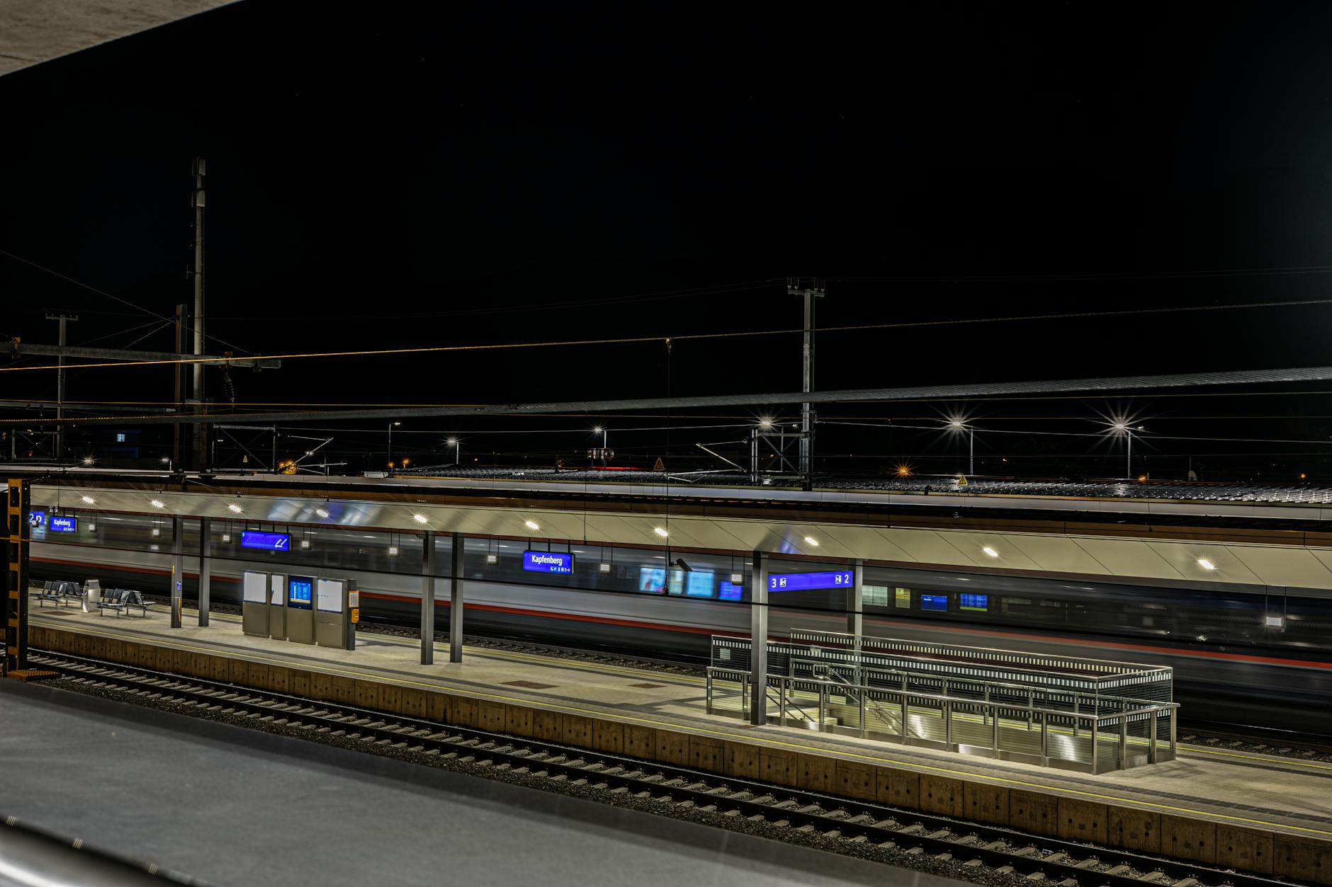 An empty railway station at night with blurred train lights passing by, showing modern infrastructure.