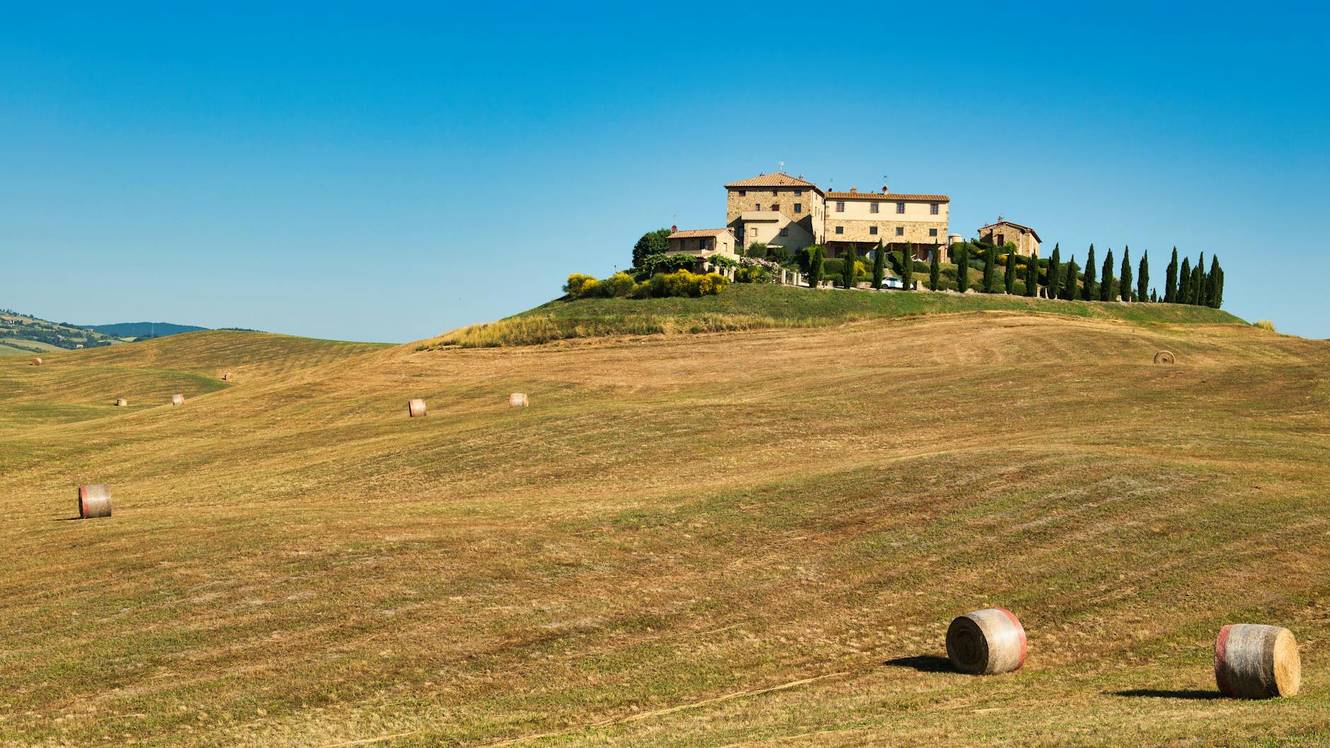 A picturesque Tuscan farmhouse atop rolling hills surrounded by hay bales under a clear summer sky.